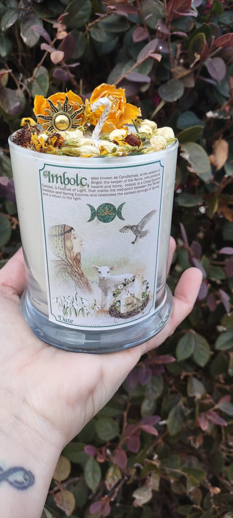 May include: A candle in a clear glass jar, decorated with dried flowers and a sun-shaped charm. The label reads "Imbolc" and features an illustration of a woman, lamb, and bird. The candle is held in a hand.