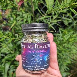 Puede incluir: Un frasco de vidrio con tapa negra lleno de hierbas secas etiquetado como "Astral Traveler Lucid Dreams". La etiqueta tambi&eacute;n incluye el texto "1 cucharadita por 6 onzas de agua hirviendo." e "Ingredientes: Ajenjo, Melisa, Damiana & Lavanda".
