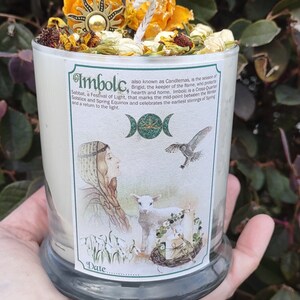 May include: A glass candle with a label featuring the text "Imbolc" and an illustration of a woman, lamb, and owl. The candle is topped with dried flowers and a sun-shaped charm.