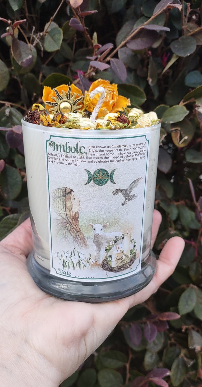May include: A glass candle with a label featuring the text "Imbolc" and an illustration of a woman, lamb, and owl. The candle is topped with dried flowers and a sun-shaped charm.