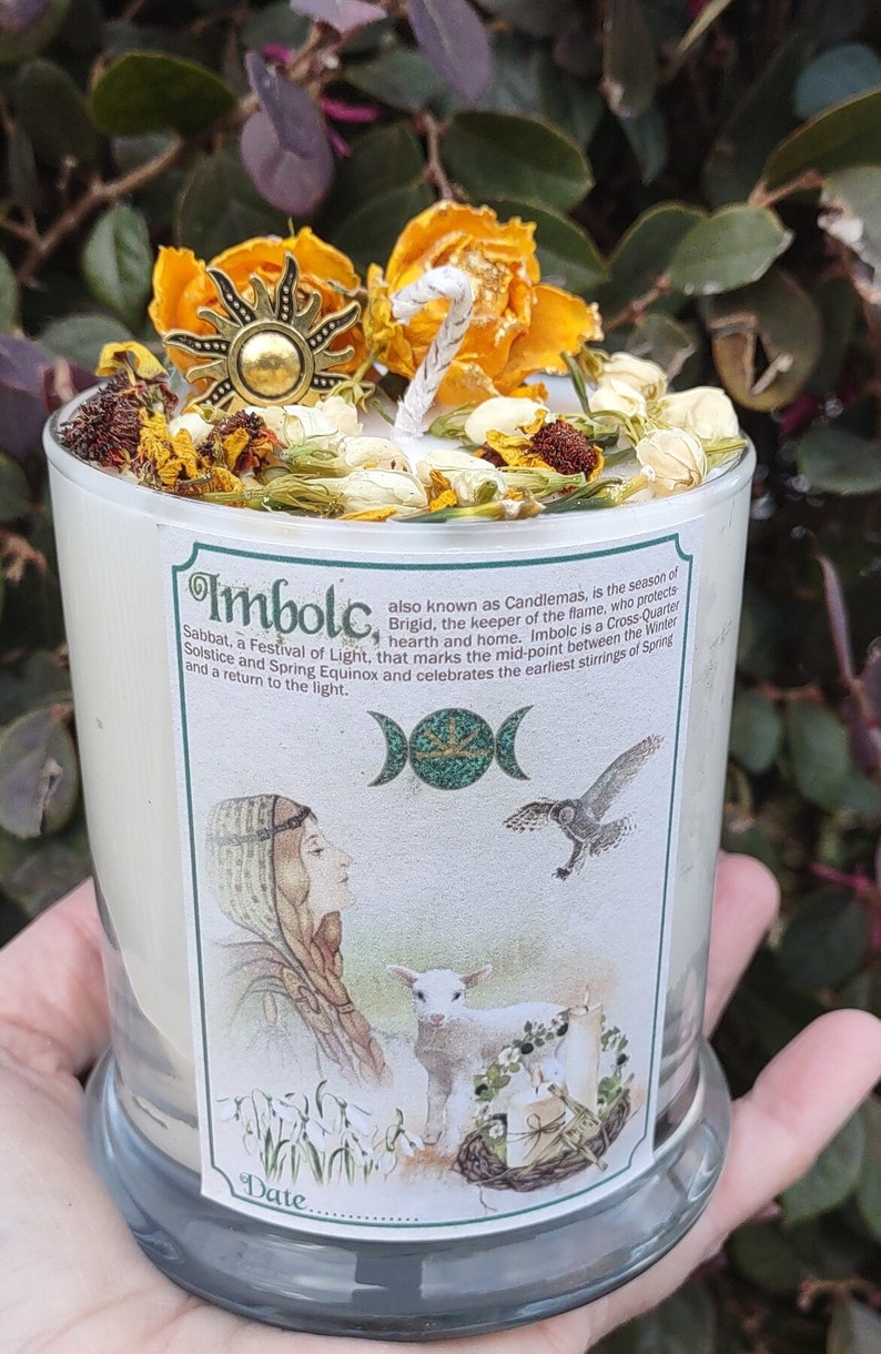May include: A glass candle with a label featuring an illustration of a woman, lamb, and owl. The candle is topped with dried flowers, a sun-shaped charm, and a white wick. The label reads "Imbolc, Sabbat, a Festival of Light."