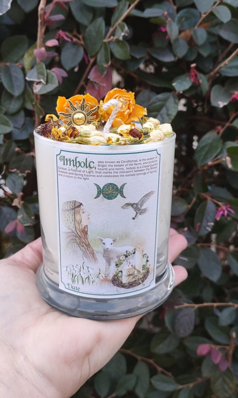 May include: A cylindrical glass candle with a label featuring illustrations and text about Imbolc. The candle is topped with dried flowers, herbs, and a sun-shaped charm. The candle is held in a hand against a backdrop of green foliage.