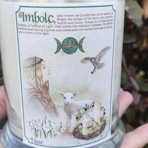 May include: A glass candle with a label featuring the word "Imbolc" and illustrations of a woman, lamb, and owl. The candle is topped with dried flowers and a sun-shaped charm. The label describes Imbolc as a festival of light.