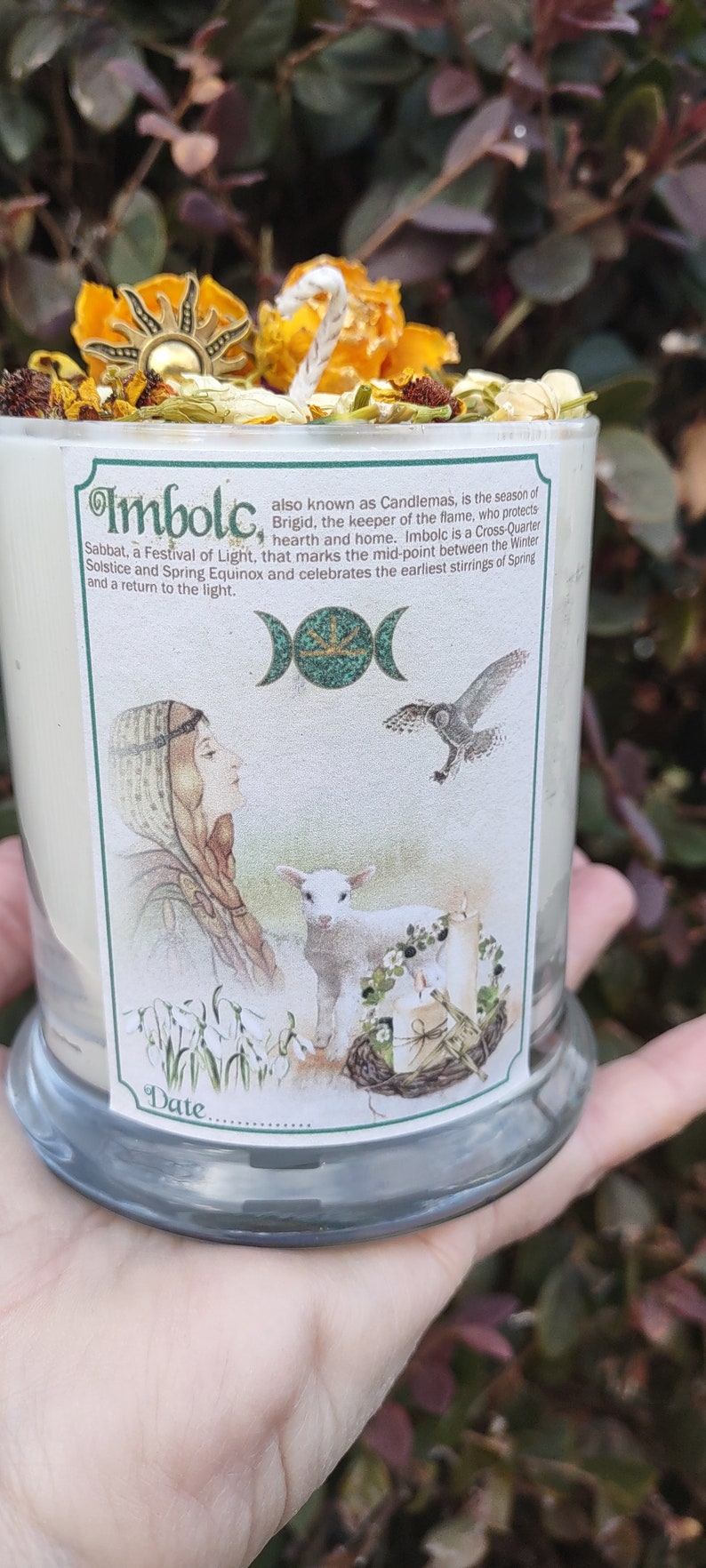 May include: A glass candle with a label featuring the word "Imbolc" and illustrations of a woman, lamb, and owl. The candle is topped with dried flowers and a sun-shaped charm. The label describes Imbolc as a festival of light.