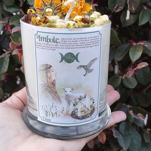 May include: A white candle in a glass jar, topped with dried flowers, a sun-shaped charm, and a white wick. The candle has a label with the text "Imbolc" and illustrations of a woman, lamb, and bird.