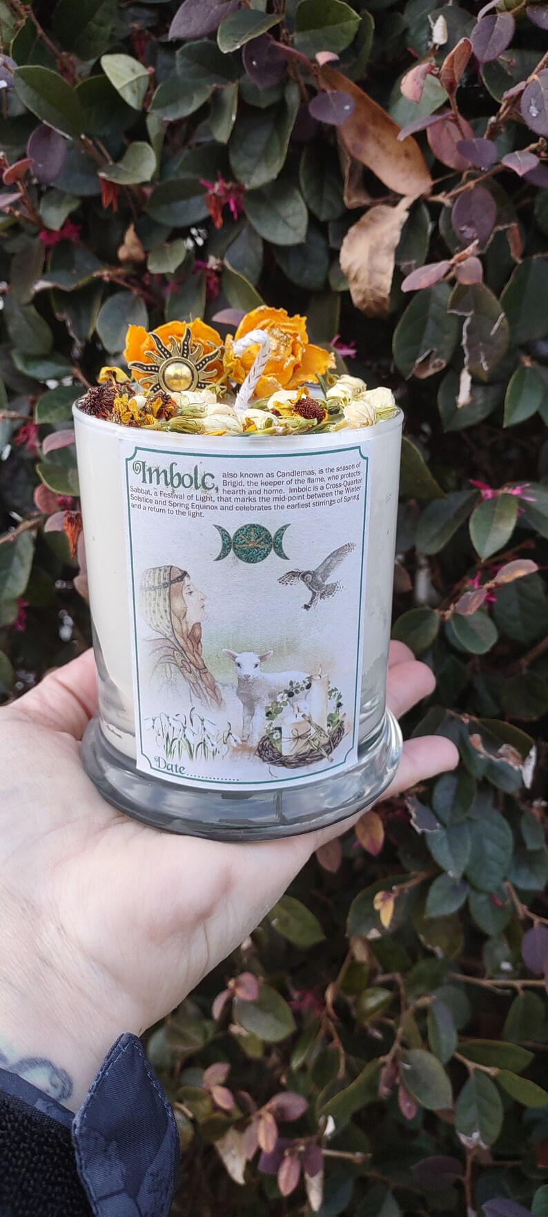 May include: A white candle in a glass jar, topped with dried flowers, a sun-shaped charm, and a white wick. The candle has a label with the text "Imbolc" and illustrations of a woman, lamb, and bird.
