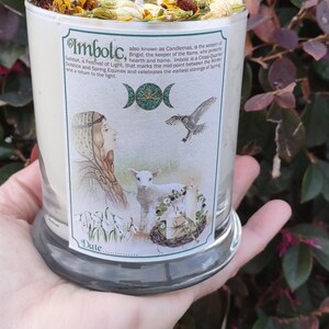 May include: A glass candle with a label featuring an illustration and the text "Imbolc". The candle is topped with dried flowers, herbs, and a sun-shaped charm. The candle is held in a hand.
