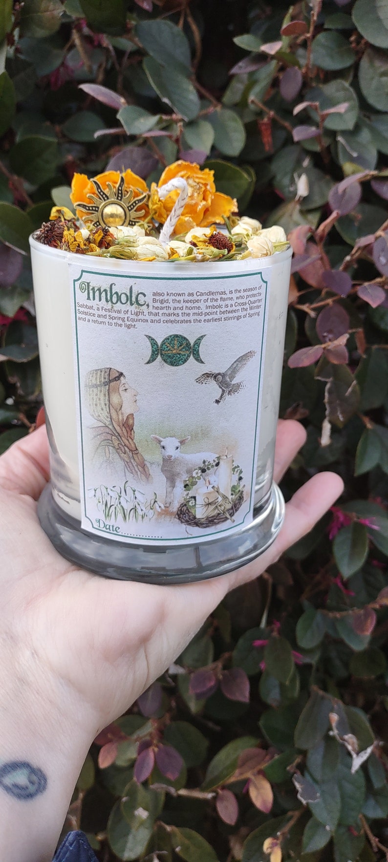 May include: A glass candle with a label featuring an illustration and the text "Imbolc". The candle is topped with dried flowers, herbs, and a sun-shaped charm. The candle is held in a hand.