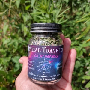 Puede incluir: Un frasco de vidrio con tapa negra lleno de hierbas secas etiquetado como "Astral Traveler Lucid Dreams". La etiqueta tambi&eacute;n incluye el texto "1 cucharadita por 6 onzas de agua. Ingredientes: Ajenjo, Melisa, Damiana & Lavanda. Ideal para la meditaci&oacute;n."