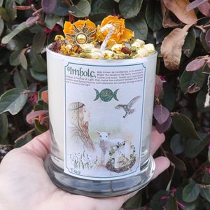 May include: A white candle with the word "Imbolc" printed on the side, topped with dried flowers and a sun-shaped charm. The candle has a label with illustrations of a woman, a lamb, and a bird. The candle is held in a person's hand.