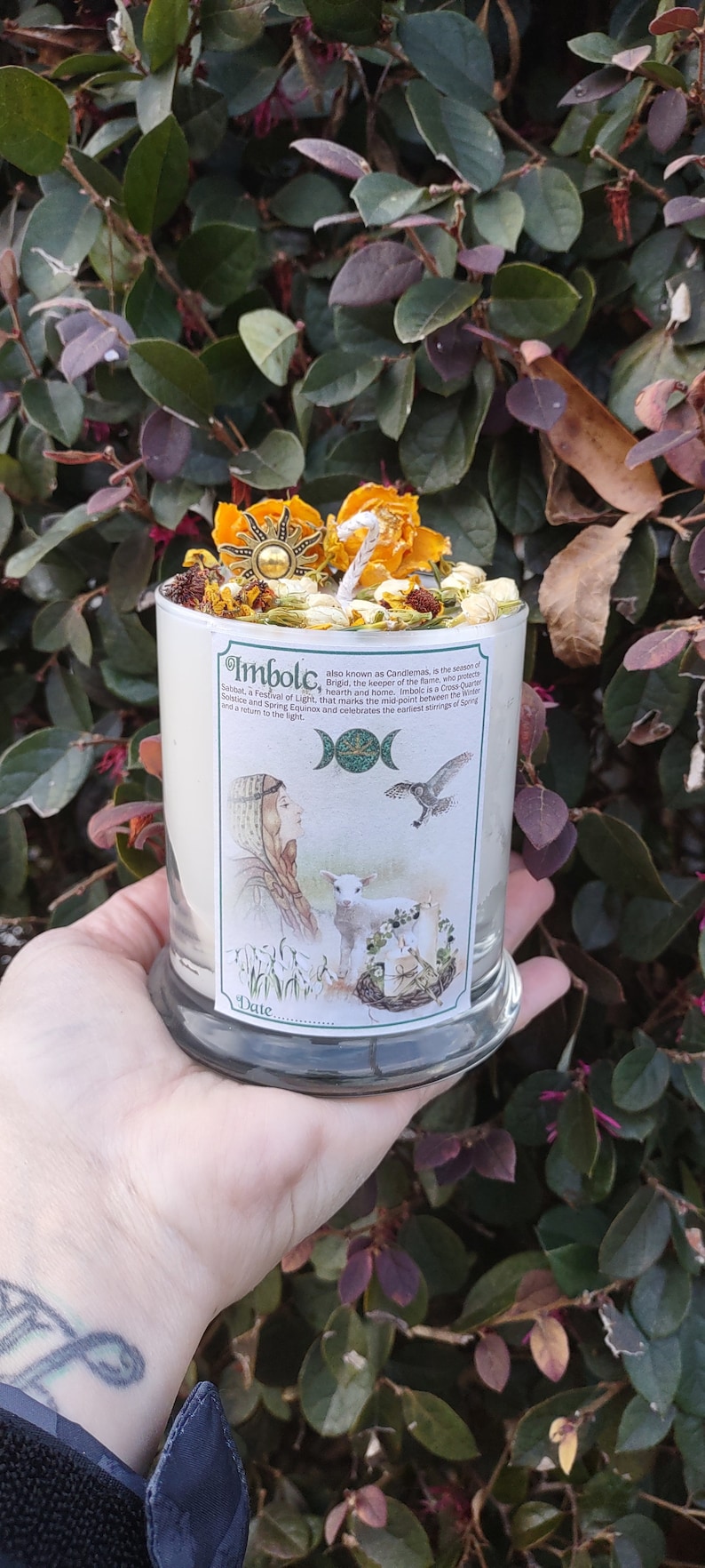 May include: A white candle with the word "Imbolc" printed on the side, topped with dried flowers and a sun-shaped charm. The candle has a label with illustrations of a woman, a lamb, and a bird. The candle is held in a person's hand.