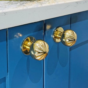 May include: Two gold cabinet knobs with a fluted design are mounted on a pair of blue cabinet doors. The cabinets are in a kitchen setting with a white countertop.