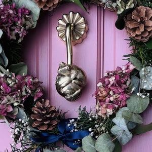 May include: A pink door with a wreath made of eucalyptus, pine cones, and pink flowers. A gold door knocker in the shape of a bird is attached to the door. A gold door handle is also visible.