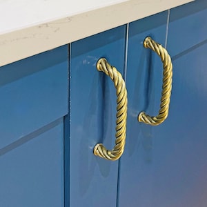 May include: Close-up of two gold rope-style cabinet pulls on a set of blue kitchen cabinets. The cabinets have a white countertop and a white tiled backsplash.
