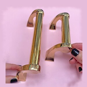 May include: Two gold-toned metal cabinet door pulls with a curved design. The pulls are different sizes.