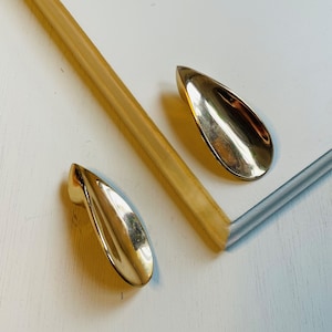 May include: Two gold-toned metal drawer pulls shaped like teardrops. The pulls are on a white surface.