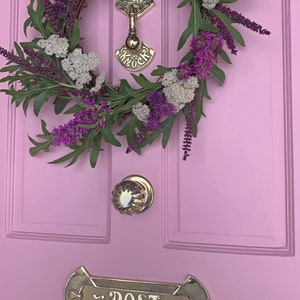 May include: A pink door with a wreath of purple and white flowers, a brass door knocker with the word "Knock" on it, a brass door knob, and a brass mailbox with the word "Post" on it.
