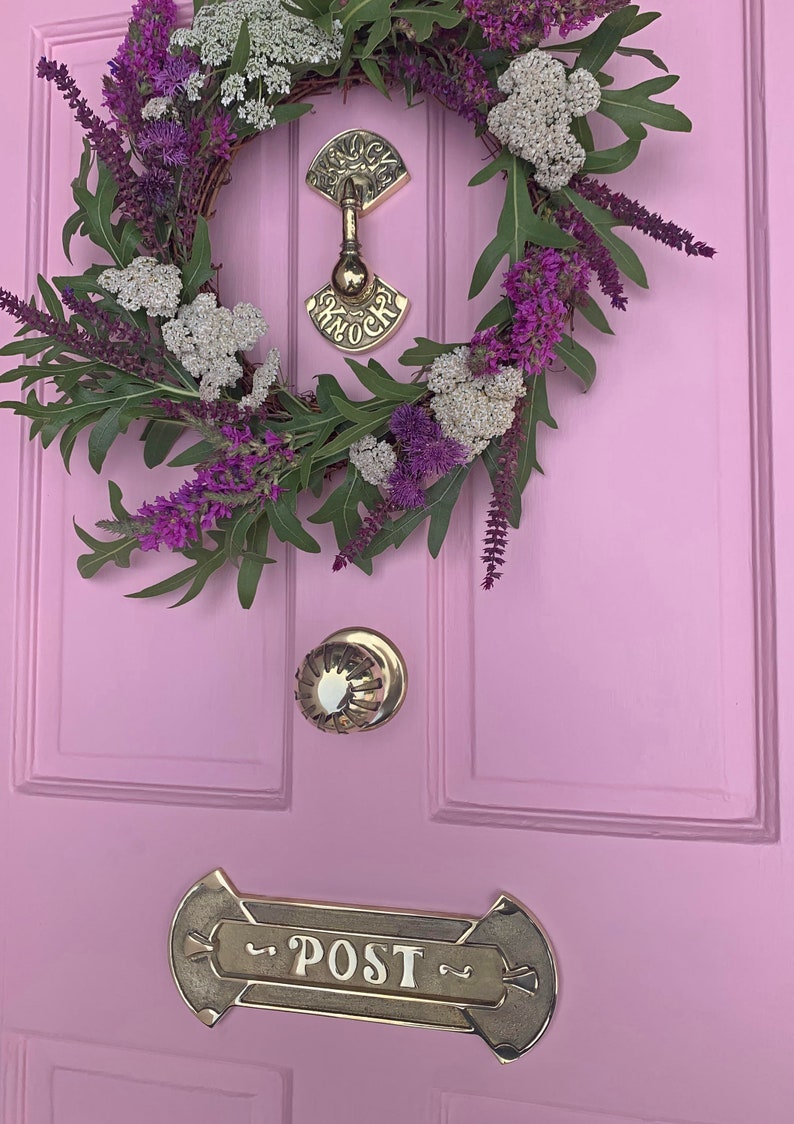 May include: A pink door with a wreath of purple and white flowers, a brass door knocker with the word "Knock" on it, a brass door knob, and a brass mailbox with the word "Post" on it.