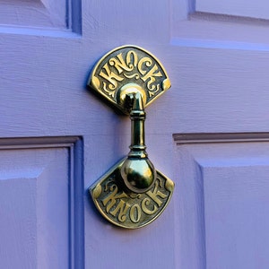 May include: A brass door knocker with a fan-shaped design and the word "Knock" inscribed on it. The knocker is attached to a purple door.