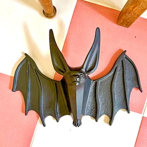 May include: A black bat-shaped decorative object with large ears and outstretched wings. The bat is made of a dark material and has a detailed face. The object is likely a Halloween or gothic-themed decoration.