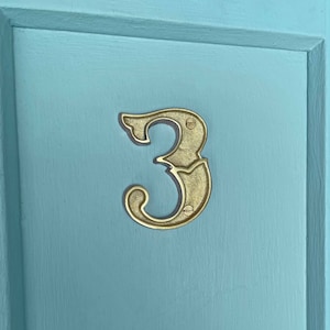 May include: A gold-colored metal house number three, with a decorative, ornate design, is attached to a light blue door.