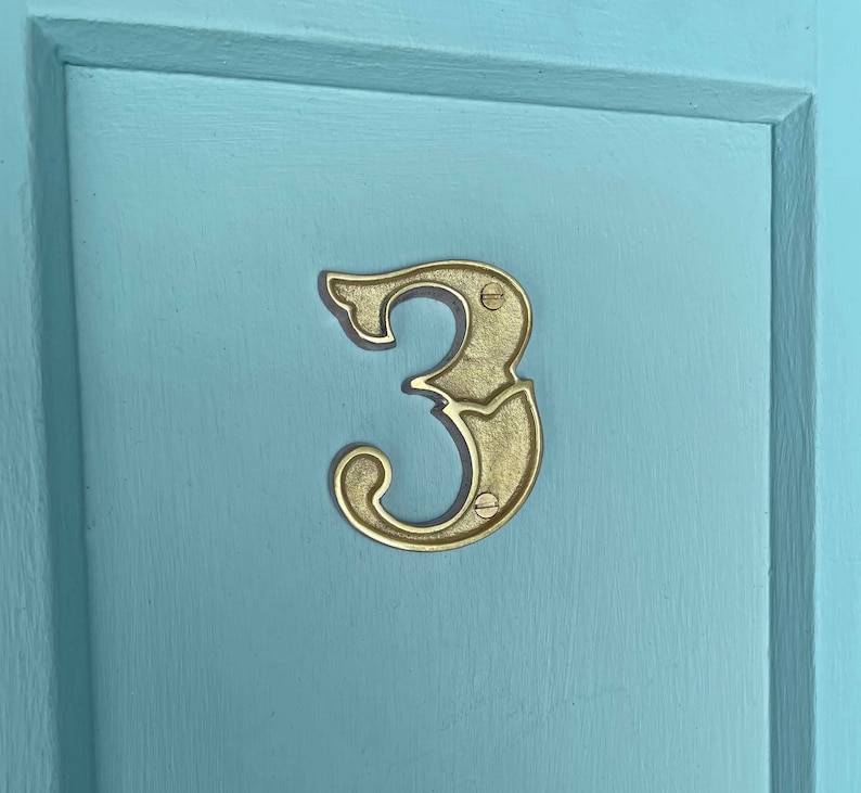 May include: A gold-colored metal house number three, with a decorative, ornate design, is attached to a light blue door.