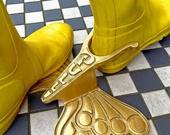 May include: A pair of bright yellow wellington boots with a gold-coloured boot jack. The boot jack has an ornate design with a curved handle and a decorative, fan-shaped base. The boots are on a black and white tiled floor.