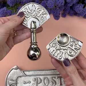 May include: A set of three silver door hardware pieces. The first piece is a door knocker shaped like a fan with the word "Knock" engraved on it. The second piece is a smaller door knocker with the word "Knock" engraved on it. The third piece is a silver door plate with the word "Post" engraved on it.