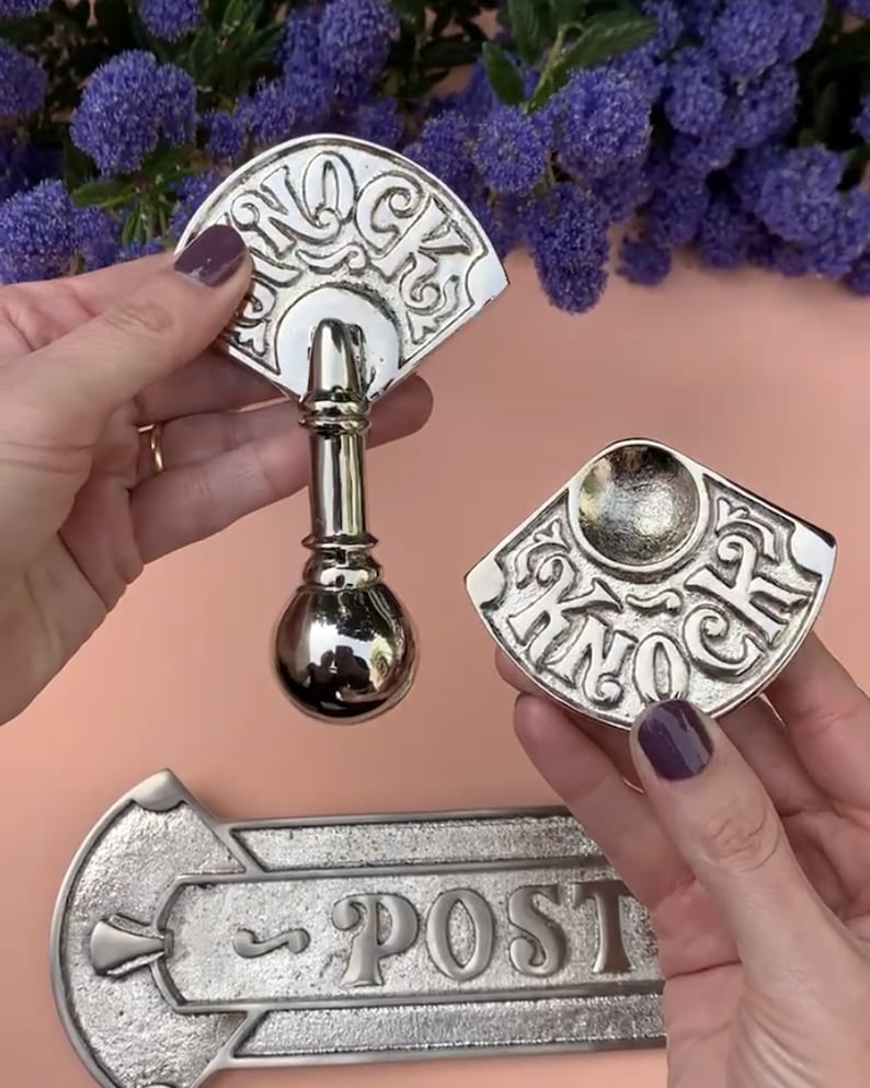 May include: A set of three silver door hardware pieces. The first piece is a door knocker shaped like a fan with the word "Knock" engraved on it. The second piece is a smaller door knocker with the word "Knock" engraved on it. The third piece is a silver door plate with the word "Post" engraved on it.