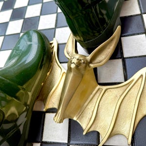 May include: A gold-colored bat-shaped boot jack with large ears and outstretched wings. The boot jack is positioned between two dark green rubber boots on a black and white checkered floor.