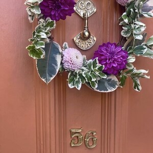 May include: A pink door with a wreath made of green leaves, white flowers, and purple flowers. The wreath is hanging on a brass door knocker that says "Go Away". The number 56 is on the door.