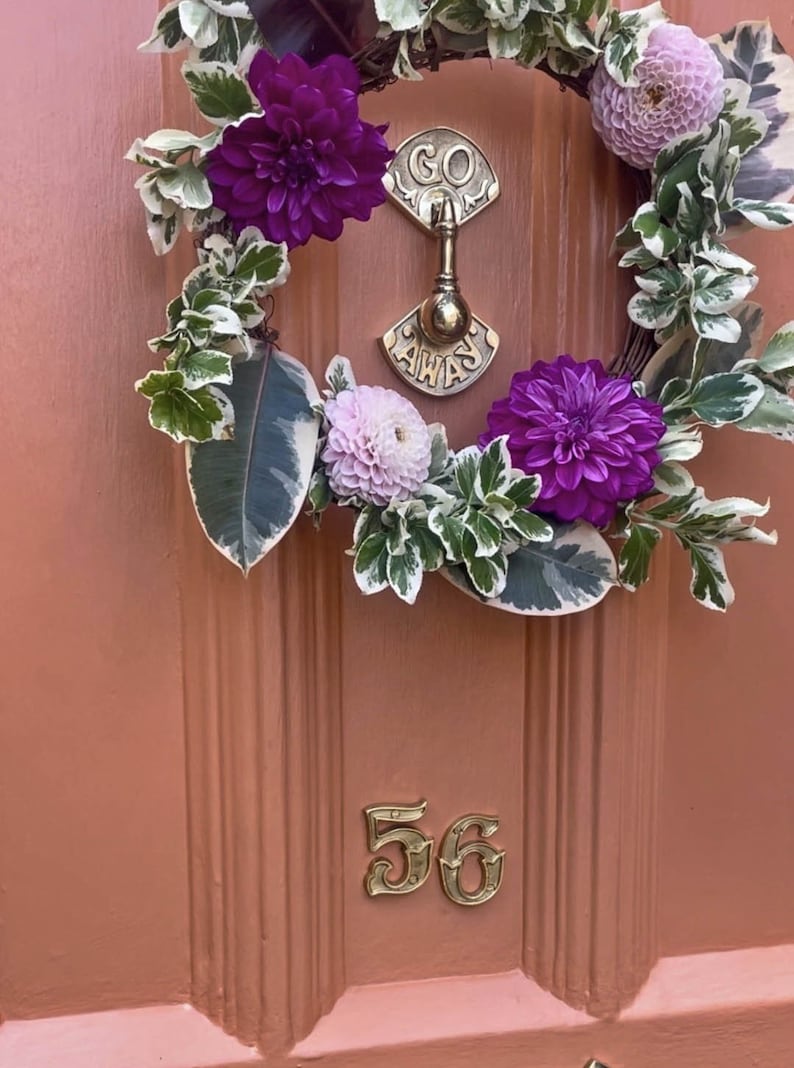 May include: A pink door with a wreath made of green leaves, white flowers, and purple flowers. The wreath is hanging on a brass door knocker that says "Go Away". The number 56 is on the door.