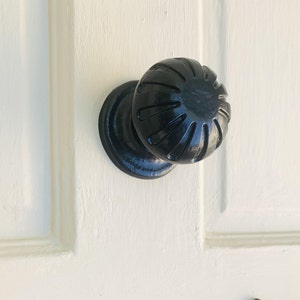 May include: A black door knob with a round, fluted design. The knob is mounted on a white door.