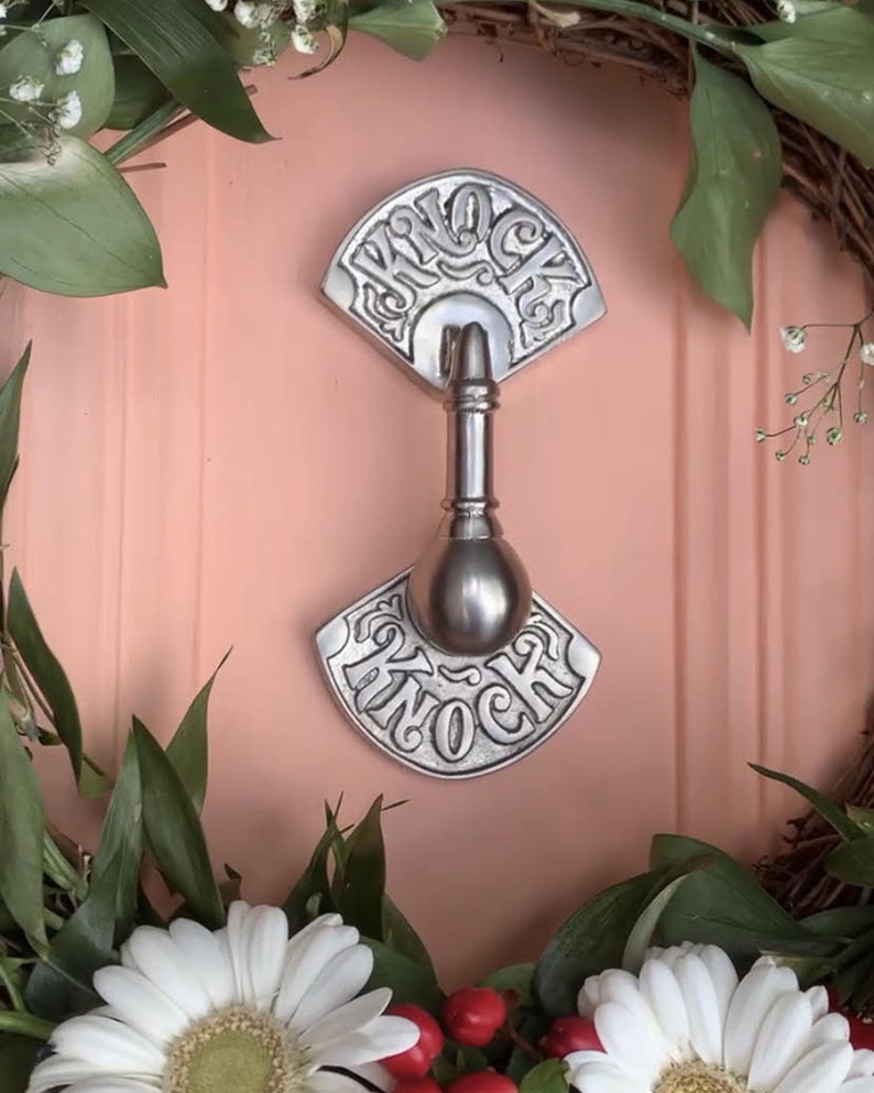 May include: A silver door knocker with the word "Knock" embossed on it. The knocker is attached to a pink door with a floral wreath around it.