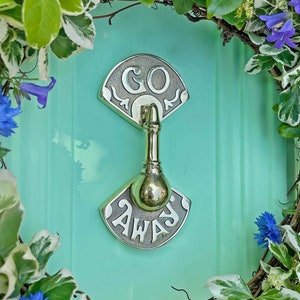 May include: A brass door knocker with a green door behind it. The knocker has two plates with the words "GO AWAY" on them.
