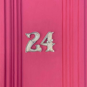 May include: A close-up of a house number, 24, on a pink door. The number is made of silver metal and is attached to the door with small screws.