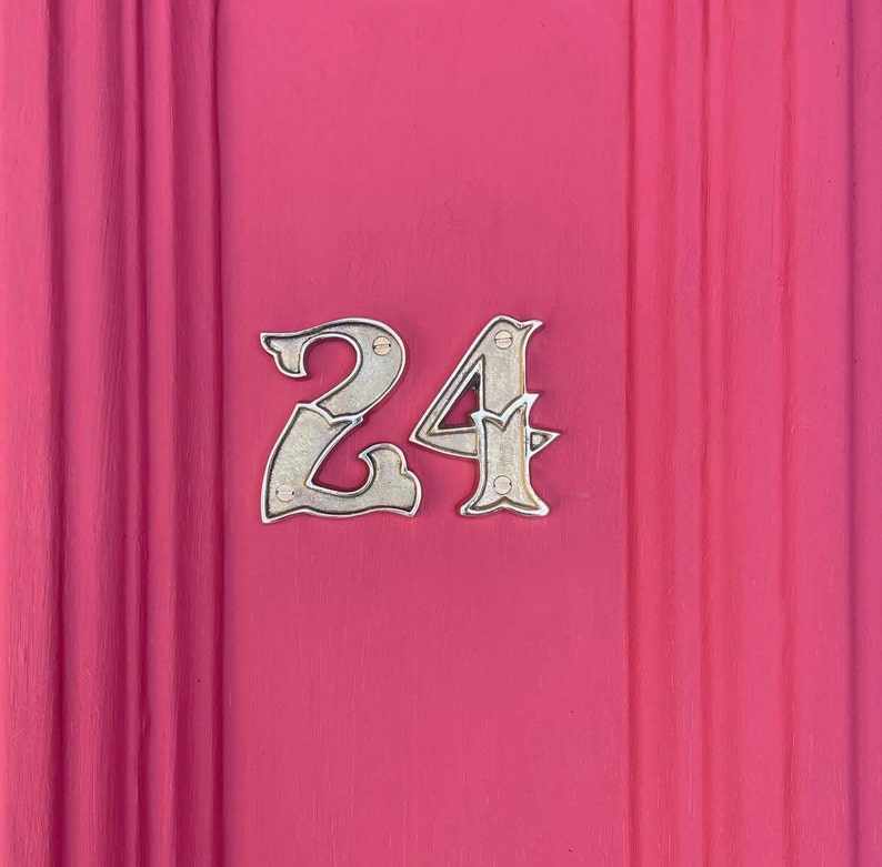 May include: A close-up of a house number, 24, on a pink door. The number is made of silver metal and is attached to the door with small screws.