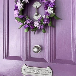 May include: A purple door with a silver door knocker and a wreath of purple and white flowers. The wreath is hanging on the door and the door knocker is in the center of the door. The door has a silver mailbox with the word "POST" on it.