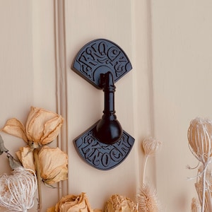May include: Black metal door knocker with a fan-shaped design and the word "Knock" inscribed on it. The knocker is attached to a wooden door.