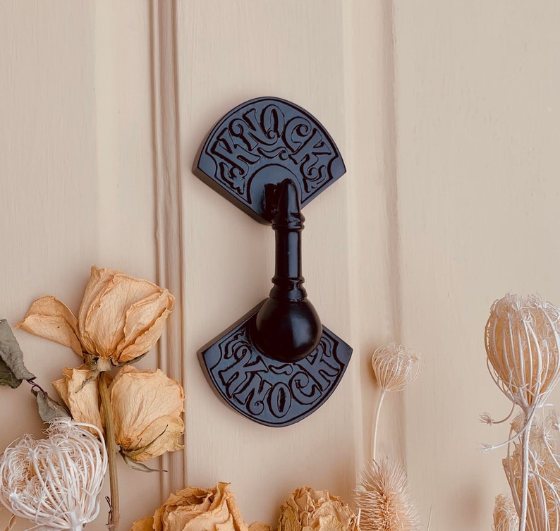 May include: Black metal door knocker with a fan-shaped design and the word "Knock" inscribed on it. The knocker is attached to a wooden door.