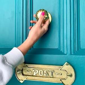May include: A hand reaches for a gold door knob on a teal blue door. Below the knob is a brass letterbox with the word "POST" engraved on it.