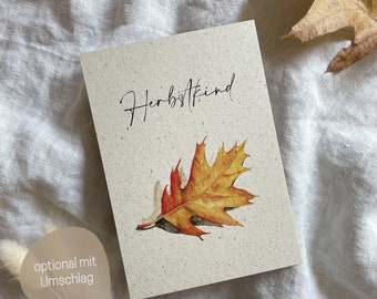 May include: A rectangular greeting card made of speckled off-white paper featuring the black cursive text "Herbstkind" above a detailed watercolor illustration of a single orange, yellow, and brown oak leaf with a visible shadow.