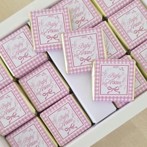 May include: A white tray containing chocolates wrapped in gold foil. Each wrapper has a pink and white gingham design with the words "Baby Atkins" inside a floral wreath and a bow.