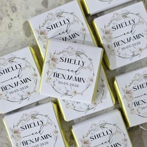 May include: Close-up of several square chocolate bars with gold foil wrappers. Each bar features a white label with the names "Shelly and Benjamin" and the date "30-05-2026" within a floral wreath. Wedding favours.