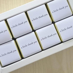 May include: A white rectangular box containing ten square chocolate pieces wrapped in gold foil. Each piece is covered by a white paper sleeve printed with the black text 'a little thank you' in a simple, clean, and legible serif font.
