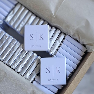 May include: A box of individually wrapped silver foil chocolates. Two chocolates are visible, each with a white label printed with the initials "S K" and the date "05.07.25". The chocolates are arranged in a cardboard box.