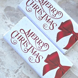 May include: Two rectangular Christmas gift boxes with white backgrounds. The boxes feature the words "MERRY CHRISTMAS" in a decorative burgundy font, along with a red bow graphic and snowflake accents. The boxes are on a light-coloured surface.