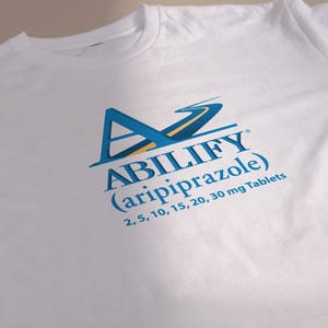 May include: White t-shirt with the word "ABILIFY" in blue and the generic name "aripiprazole" below it. The shirt also displays the tablet strengths: 2, 5, 10, 15, 20, and 30 mg. The shirt has a crew neck.