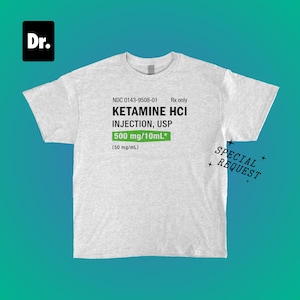 Puede incluir: Camiseta gris claro con el texto "KETAMINE HCI INJECTION, USP 500 mg/10mL*" en negro y verde. La camiseta también tiene el texto "SPECIAL REQUEST" en negro. Un cuadrado negro con "Dr." en blanco está en la esquina superior izquierda.