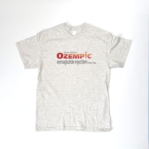 以下が含まれることがあります： 赤と黒で「ONCE-WEEKLY OZEMPIC semaglutide injection 0.5mg 1mg」と書かれた、ライトグレーの半袖Tシャツ。白い面に平らに置かれています。